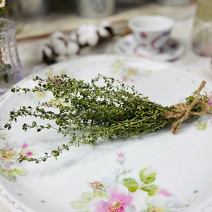 May include: A bunch of green herbs tied with twine, resting on a white plate with a floral design. The plate has a light blue rim and features pink roses.