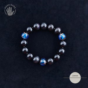Könnte beinhalten: Handgefertigtes Armband mit schwarzen Perlen und drei blauen und goldenen Akzentperlen. Das Armband wird auf dunklem Hintergrund gezeigt. Das Bild enthält den Text "HAND MADE" und den Markennamen "AJKTN".