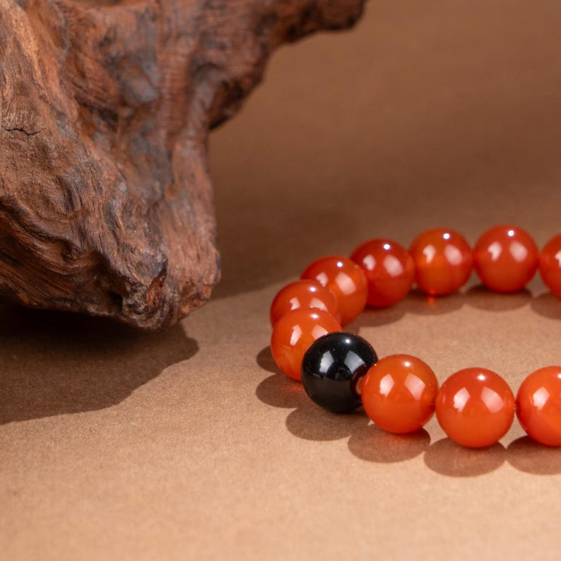 May include: A beaded bracelet featuring orange and black spherical beads. The bracelet is made of polished, round beads in a vibrant orange color, with one contrasting black bead. The bracelet is displayed on a brown surface.