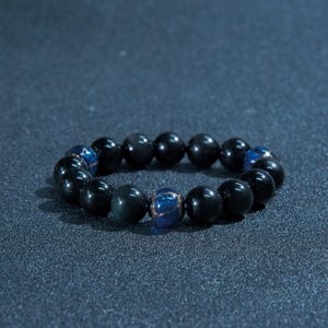 May include: A black beaded bracelet featuring a mix of black and blue beads. The bracelet is made of round, polished beads. The blue beads have a speckled appearance. The bracelet is displayed on a dark blue surface.