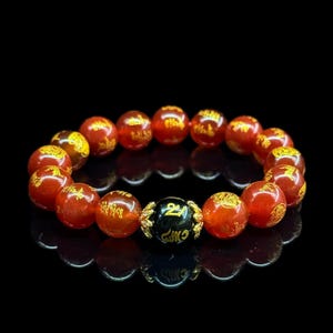 May include: A red beaded bracelet with gold-colored inscriptions. The bracelet features a black bead with gold-colored symbols. The beads are round and have a glossy finish. The bracelet is displayed on a black surface.