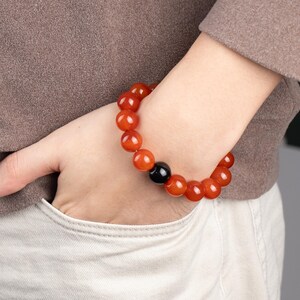 May include: A beaded bracelet featuring large, round, reddish-orange beads. A single black bead is interspersed among the orange beads. The bracelet is worn on a wrist, with the hand resting in a pocket.