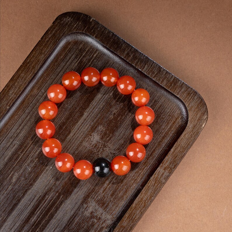 May include: A bracelet featuring large, round, orange beads with a glossy finish. The bracelet includes a single, contrasting black bead. The beads are strung together, forming a circular shape, and are displayed on a wooden tray.
