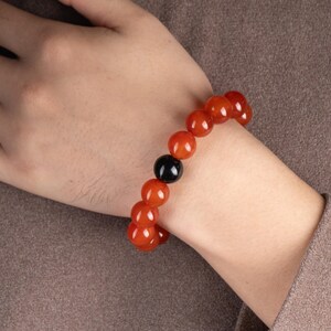 May include: A beaded bracelet featuring large, round, reddish-orange beads interspersed with a single black bead. The bracelet is worn on a wrist, showcasing the contrast between the colors and textures.