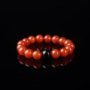 May include: A beaded bracelet featuring orange-red, round beads with a single, black, round bead. The bracelet is displayed against a black background, with a reflective surface.