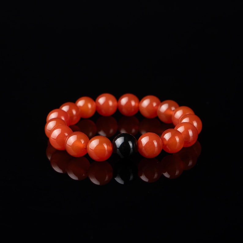 May include: A beaded bracelet featuring orange-red, round beads with a single, black, round bead. The bracelet is displayed against a black background, with a reflective surface.