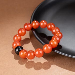 May include: A beaded bracelet featuring large, round, orange beads with a glossy finish. One black bead is interspersed among the orange beads. The bracelet is resting on a dark gray surface, with a brown background.