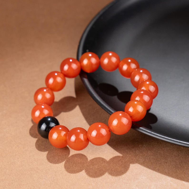 May include: A beaded bracelet featuring large, round, orange beads with a glossy finish. One black bead is interspersed among the orange beads. The bracelet is resting on a dark gray surface, with a brown background.