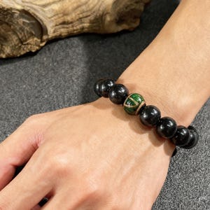 Könnte beinhalten: Schwarzes Perlenarmband mit einer einzelnen grünen und goldenen Akzentperle. Das Armband besteht aus glatten, runden, schwarzen Perlen. Die Akzentperle hat ein grünes Design mit goldenen Akzenten. Das Armband wird am Handgelenk getragen.