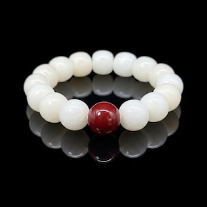 May include: A white beaded bracelet featuring a single, large, deep red bead. The bracelet is composed of round, smooth beads and is set against a black background, creating a reflective surface.