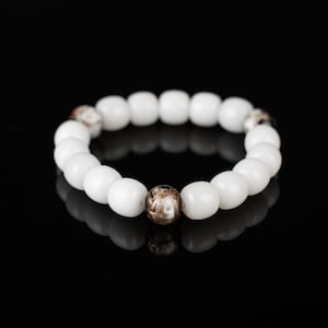 May include: White beaded bracelet with a single brown and gold accent bead. The bracelet is made of round, glossy white beads and is displayed against a black background.