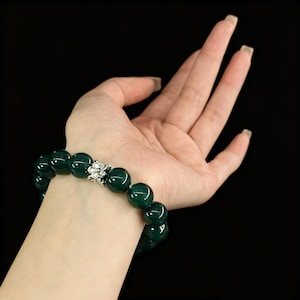 Puede incluir: Una pulsera de cuentas verde oscuro con un acento plateado en forma de flor. La pulsera está hecha de cuentas grandes y redondas. La pulsera está en una muñeca sobre un fondo negro.