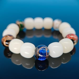 Peut inclure: Bracelet de perles composé de perles rondes blanc cassé et de perles de verre décoratives. Les perles de verre sont bleues, dorées, rouges et vertes. Le bracelet est présenté sur un fond bleu réfléchissant. Les perles mesurent environ 1 cm de diamètre.