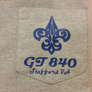 May include: A gray t-shirt with a blue pocket featuring a stylized fleur-de-lis design and the text "GT 840 Stafford VA".