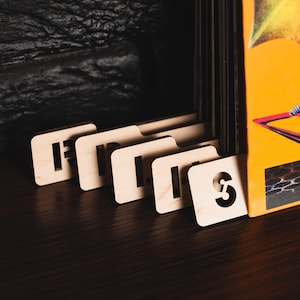 May include: Wooden record dividers with cut-out letters and numbers, including the number 3. The dividers are placed in front of a stack of vinyl records and a colourful album cover. The dividers are on a dark wooden surface.