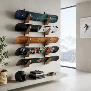 May include: A wall-mounted wooden rack holding five snowboards of varying colors and designs, including black, white, and wood-grain finishes. Below the rack sits a black helmet, a stack of folded clothing, and a coiled rope on a concrete ledge.