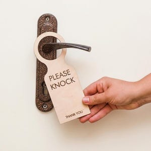Może przedstawiać: Drewniany wieszak na drzwi z nadrukiem "PLEASE KNOCK" i "THANK YOU". Wieszak na drzwi wisi na brązowym uchwycie drzwi. Tło to jasne, kremowe drzwi.