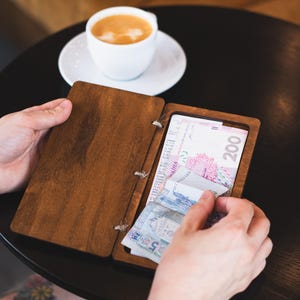 Op de afbeelding: Een houten bonnenhouder met een donkerbruine afwerking, open om een ​​bon en Oekraïense valuta te onthullen. De houder wordt vastgehouden door de handen van een persoon. Een witte kop koffie staat op een schotel op de achtergrond.