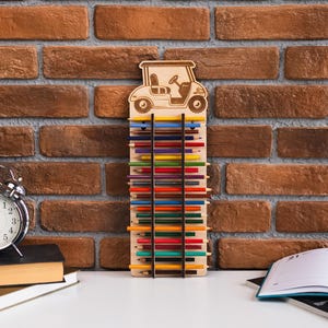 Peut inclure: Porte-crayons en bois en forme de voiturette de golf, rempli de crayons de couleur. Le support est en bois clair et présente un motif de voiturette de golf en haut. Les crayons sont disposés verticalement, affichant une variété de couleurs.