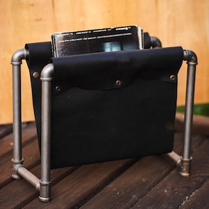 Puede incluir: Un revistero de cuero negro y metal. El revistero tiene forma rectangular con un bolsillo de cuero negro y está sostenido por un marco de tubería metálica. El revistero está sobre una superficie de madera.