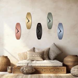 May include: A collection of six oval-shaped wall clocks in various colors: copper, gold, black, sage green, and blue. Each clock has black hands and the number 12 at the top. The clocks are mounted on a neutral-colored wall.