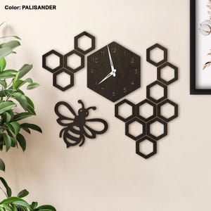 May include: A dark brown wooden wall clock with a honeycomb design and a bee silhouette. The clock has white hands. The colour is listed as Palisander. Additional honeycomb shapes surround the clock.
