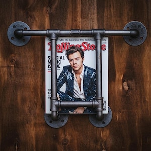 Puede incluir: Un revistero de estilo industrial hecho de tuberías negras, montado sobre una superficie de madera. El estante contiene una revista con el logotipo de Rolling Stone y una foto de una persona. El estante está fijado a la pared con placas metálicas circulares.
