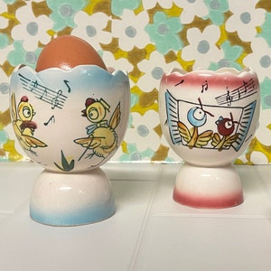 May include: Two white ceramic egg cups with blue and pink accents. Each cup features a whimsical design of birds singing with musical notes. One cup has a brown egg inside.