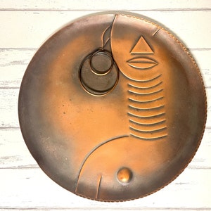 May include: A round copper tray with a stylized design of a face with a geometric pattern. The tray has a hammered texture and a dark brown patina.