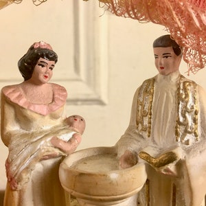 Puede incluir: Un conjunto de figuritas de cerámica vintage que representa una ceremonia de bautismo. Una mujer con un vestido blanco sostiene un bebé en sus brazos, mientras que un sacerdote con una túnica blanca con detalles dorados se encuentra junto a una pila bautismal.