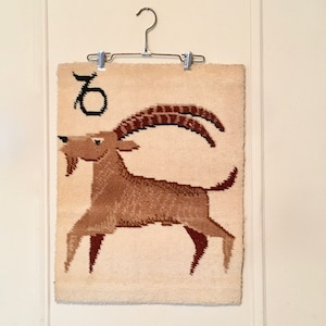 May include: A beige woven wall hanging with a brown and black image of a goat with a fish tail and the astrological symbol for Capricorn.