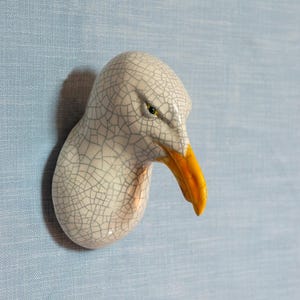 May include: A ceramic seagull head wall sculpture with a white crackled glaze and a yellow beak. The seagull's eye is painted a bright yellow.