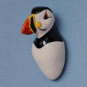Puede incluir: Una escultura de pared de cerámica con forma de frailecillo, que presenta una cabeza y espalda de color negro brillante, un pecho blanco con un acabado de esmalte craquelado fino y un pico de colores vibrantes en naranja, amarillo y negro.