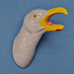 Puede incluir: Una escultura de pared de cerámica con forma de cabeza de gaviota y acabado de esmalte blanco craquelado. El ave presenta un pico amarillo brillante, la boca abierta mostrando una lengua rosa, una mancha roja y un ojo oscuro con iris amarillo.