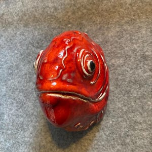 Puede incluir: Un recipiente de cerámica con forma de pez, con un esmalte rojo brillante y vibrante. El pez tiene detalles, incluyendo ojos, aletas y boca. El recipiente se utiliza probablemente con fines decorativos o funcionales.