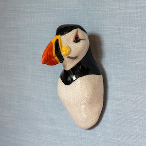 May include: A ceramic sculpture of a puffin with a black and white head and a bright orange and yellow beak. The puffin is facing to the left and has a white body with a cracked glaze.