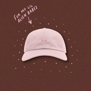 May include: Pink baseball cap with an alien face embroidered on the front. The text "For my lil alien babes" is written above the cap.
