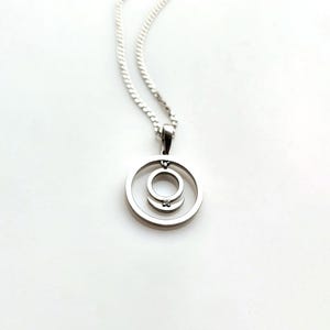 Op de afbeelding: Een zilveren hangerketting met een dubbele cirkel ontwerp. De hanger heeft een kleine, heldere edelsteen in het midden van de binnenste cirkel. De ketting is ook zilverkleurig en wordt tegen een witte achtergrond getoond.
