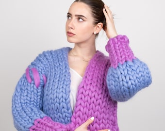 May include: A woman wearing a blue and purple chunky knit cardigan with a white top and blue jeans.