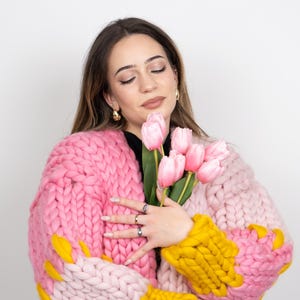 Può includere: Una donna indossa un cardigan di maglia grossa rosa e giallo con maniche lunghe e una vestibilità rilassata. Tiene un mazzo di tulipani rosa.