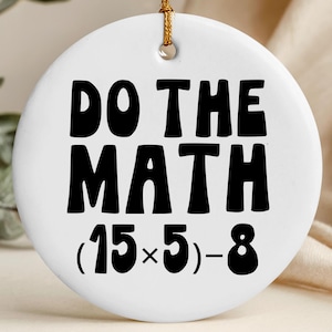 May include: A white, round ceramic ornament with the phrase "DO THE MATH" in bold, black letters. Below the text is the equation (15x5)-8. A gold string is attached for hanging. The ornament is a decorative item.