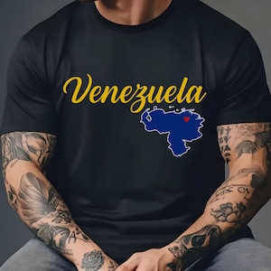 May include: Black t-shirt featuring the word "Venezuela" in yellow script above a blue map of the country with a red heart. The shirt is a crew neck and is being worn.