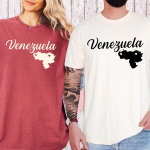 May include: Two t-shirts, one red and one cream, each with the word "Venezuela" in cursive script. Below the word is a map of Venezuela with a heart. The red shirt has a rolled-up sleeve. The cream shirt has a short sleeve.