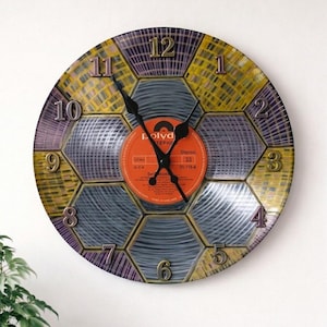 Puede incluir: Un reloj de pared decorativo con un centro de vinilo vintage. La esfera del reloj presenta un diseño de panal en tonos amarillos, morados y grises. Las manecillas del reloj son negras y los números metálicos.