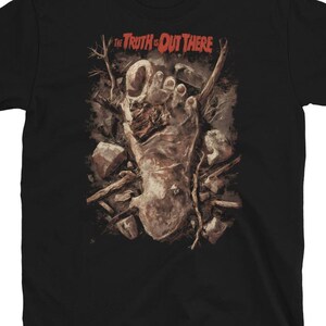 Pode incluir: T-shirt preto com uma ilustração gráfica de um pé em decomposição rodeado por rochas e galhos. O texto "The Truth Is Out There" é impresso acima do pé em vermelho e branco. A camisa é feita de um material macio.