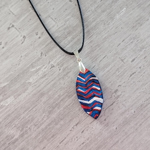 May include: A handmade pendant necklace with a black cord. The pendant is an elongated oval shape with a silver bail. The pendant features a chevron pattern in red, white, and blue. The necklace is displayed on a gray surface.