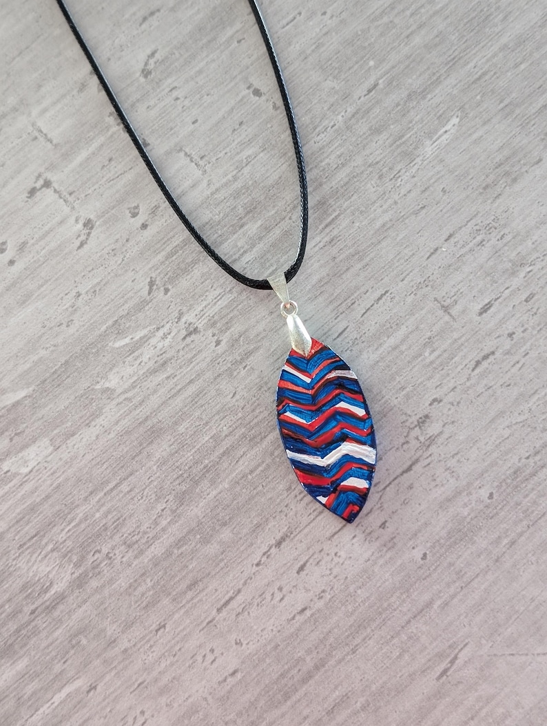 May include: A handmade pendant necklace with a black cord. The pendant is an elongated oval shape with a silver bail. The pendant features a chevron pattern in red, white, and blue. The necklace is displayed on a gray surface.
