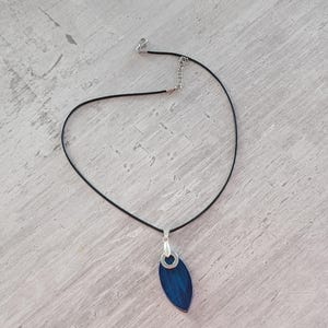 May include: A pendant necklace featuring a dark blue, leaf-shaped pendant with a silver-colored bail. The pendant hangs from a thin, black cord necklace with a silver-colored clasp and adjustable chain.