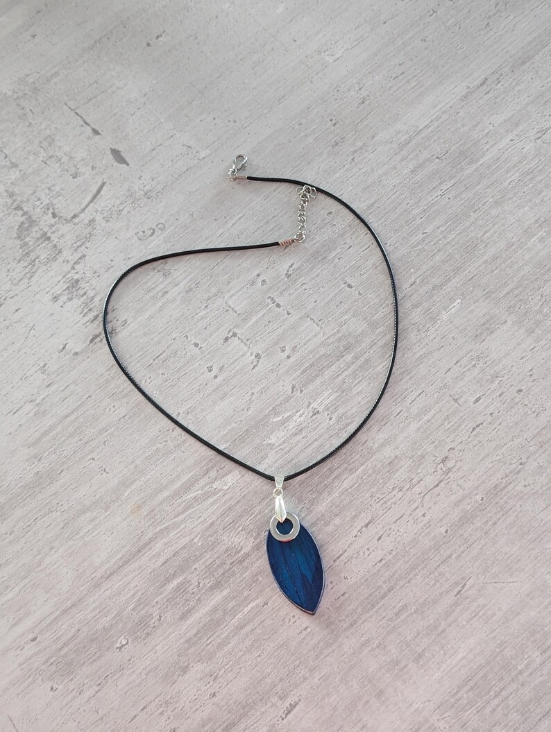 May include: A pendant necklace featuring a dark blue, leaf-shaped pendant with a silver-colored bail. The pendant hangs from a thin, black cord necklace with a silver-colored clasp and adjustable chain.