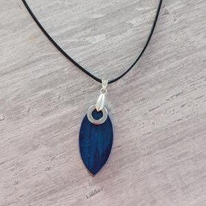 May include: A pendant necklace featuring a dark blue, leaf-shaped pendant with a silver-colored metal border and a silver-colored metal loop. The pendant hangs from a thin, black cord. The necklace is displayed on a light gray surface.